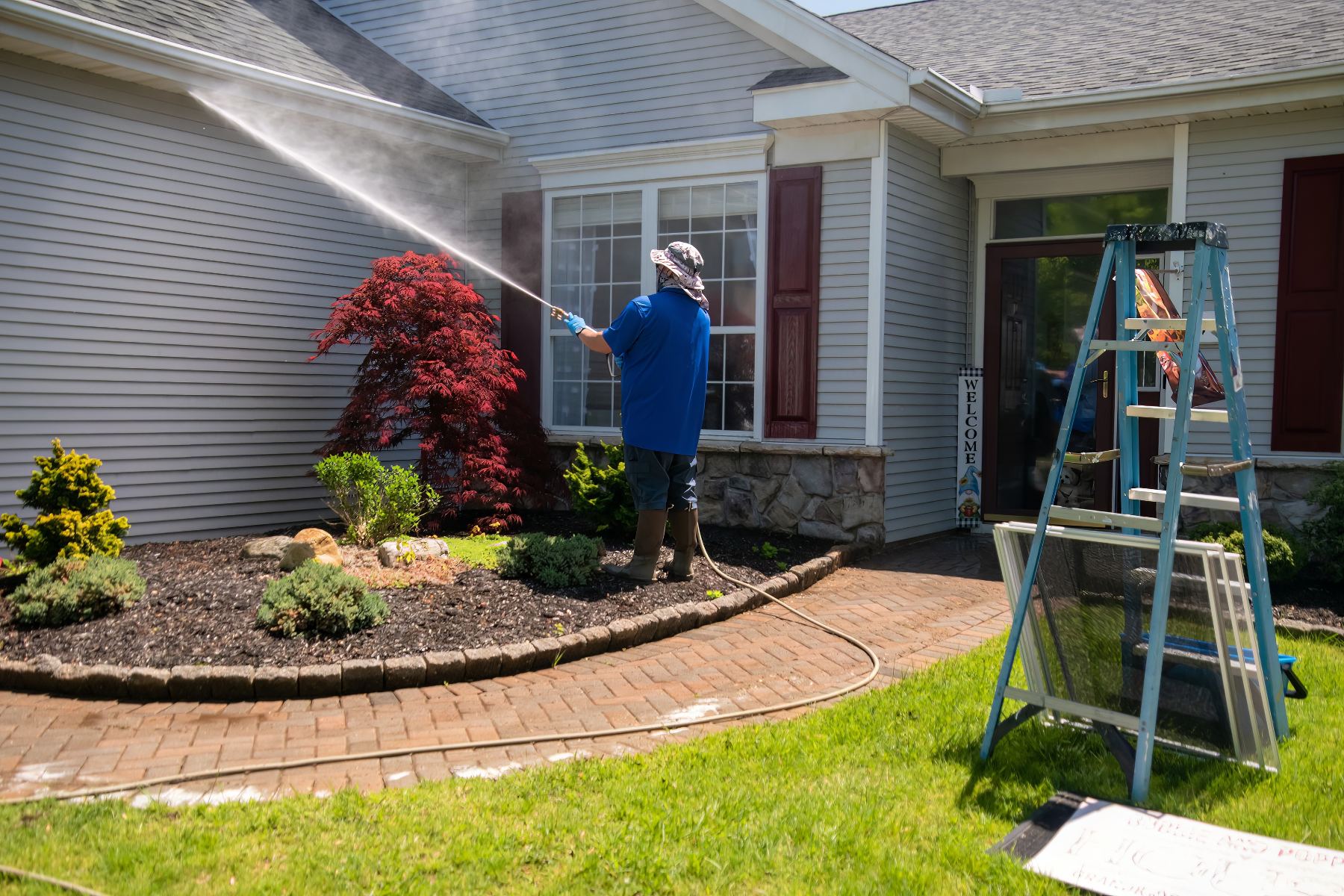 Man in blue jacket pressure washing house siding with ladder nearby